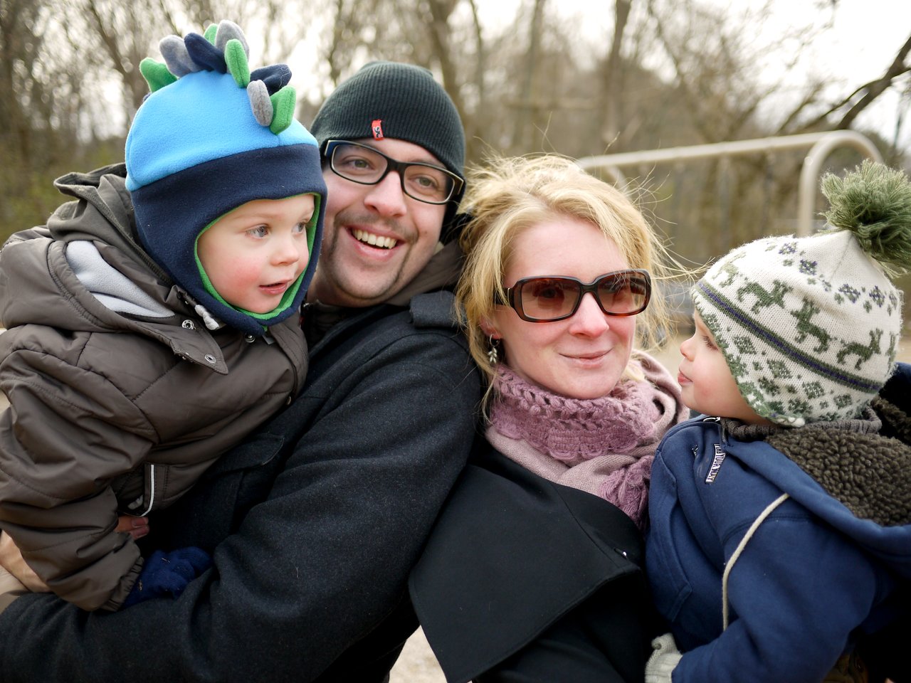 A man and woman stand close together, each holding a young child dressed in warm winter clothing.