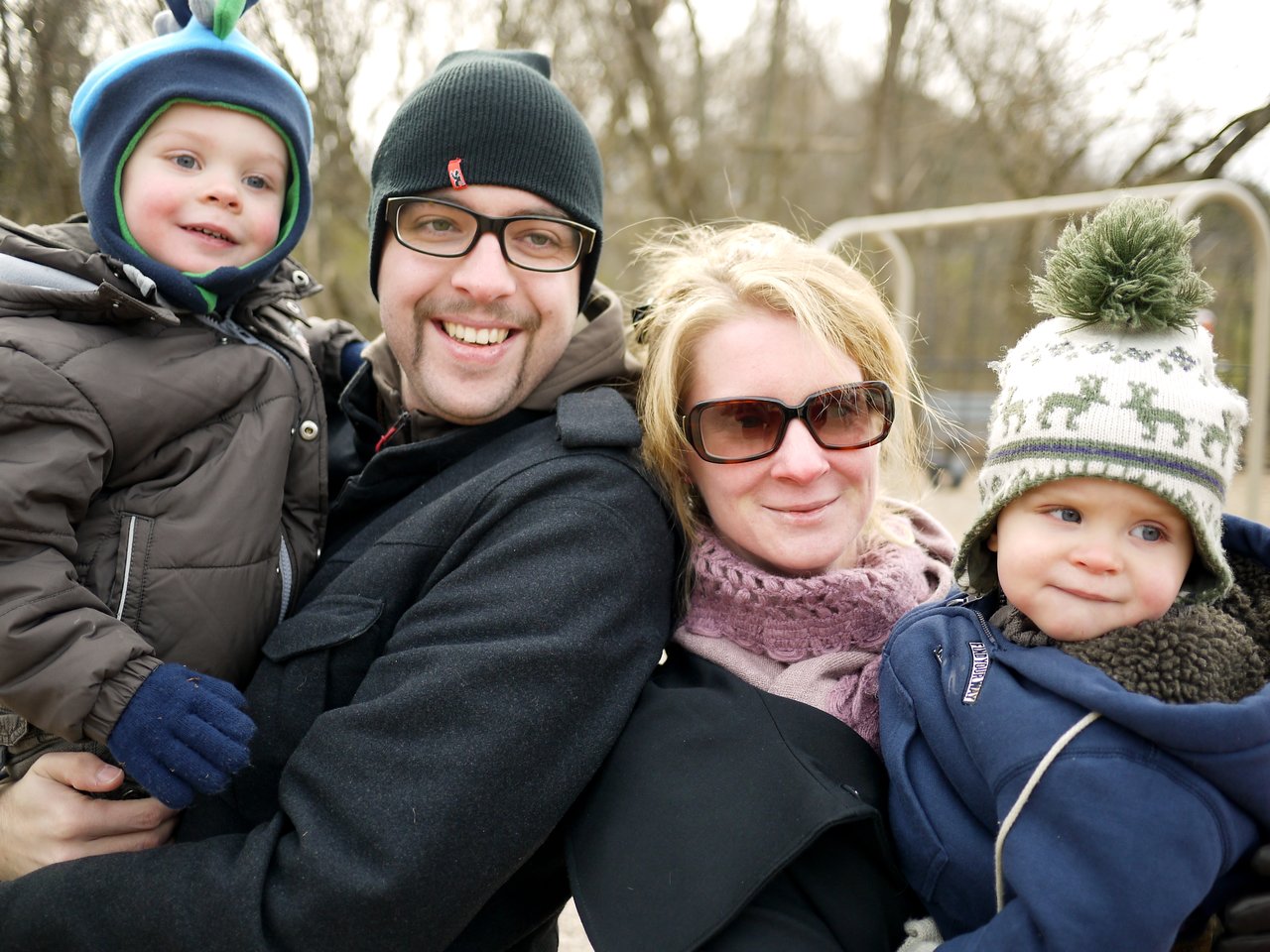A man and woman stand close together, each holding a young child dressed in warm winter clothing.