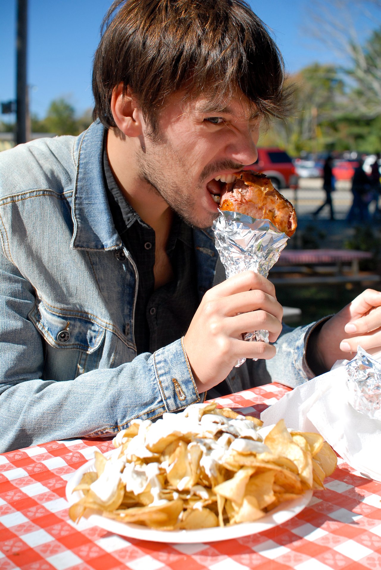 A man in a denim jacket bites into a large roasted meat piece wrapped in foil at an outdoor table.