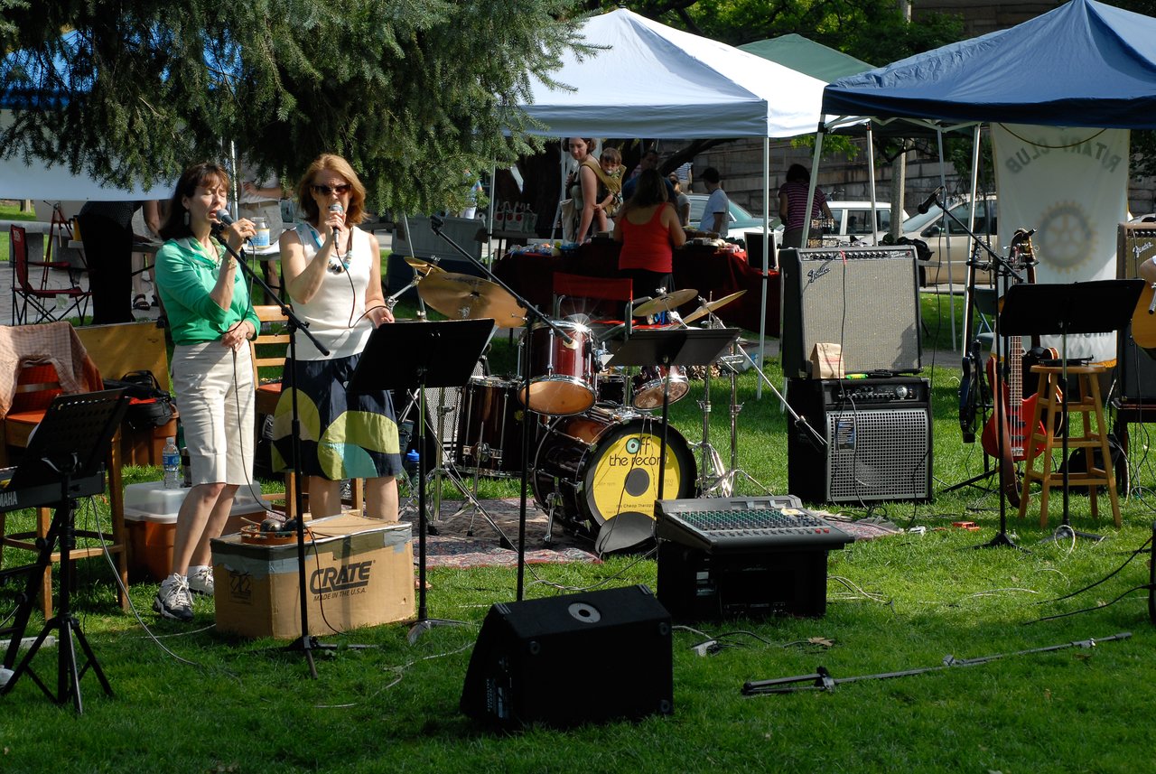 Two women sing into microphones at an outdoor farmers market, with a live band setup and tents in the background.