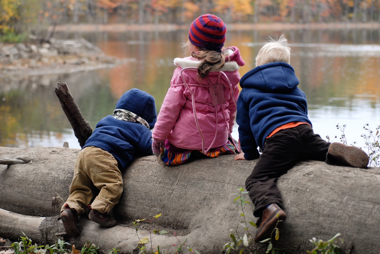 Three children in warm clothing sit and lean on a fallen log, looking at a calm lake in autumn.
