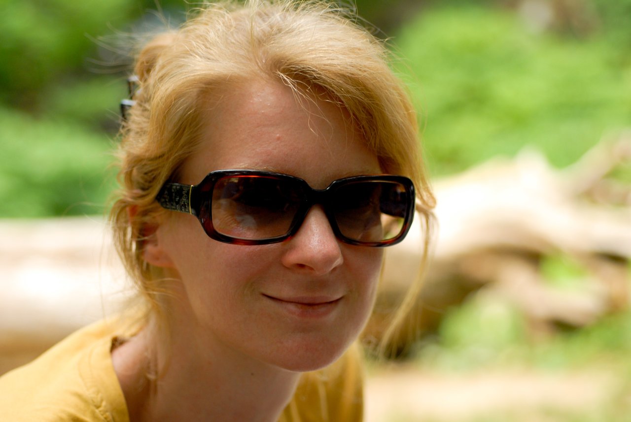 A woman with blonde hair and dark sunglasses smiles slightly while looking at the camera in an outdoor setting.