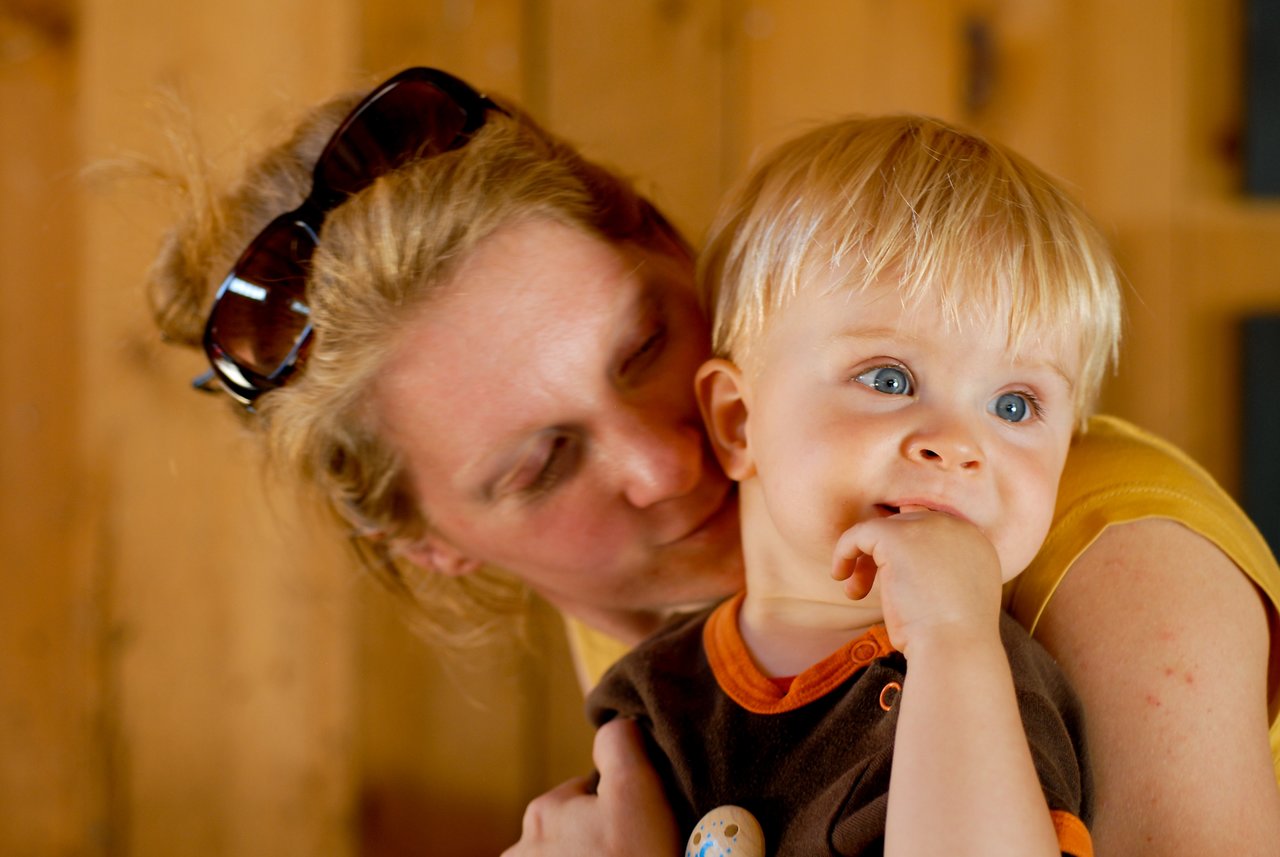 A woman holds a young child close, gently leaning in while the child looks away with a finger in their mouth.