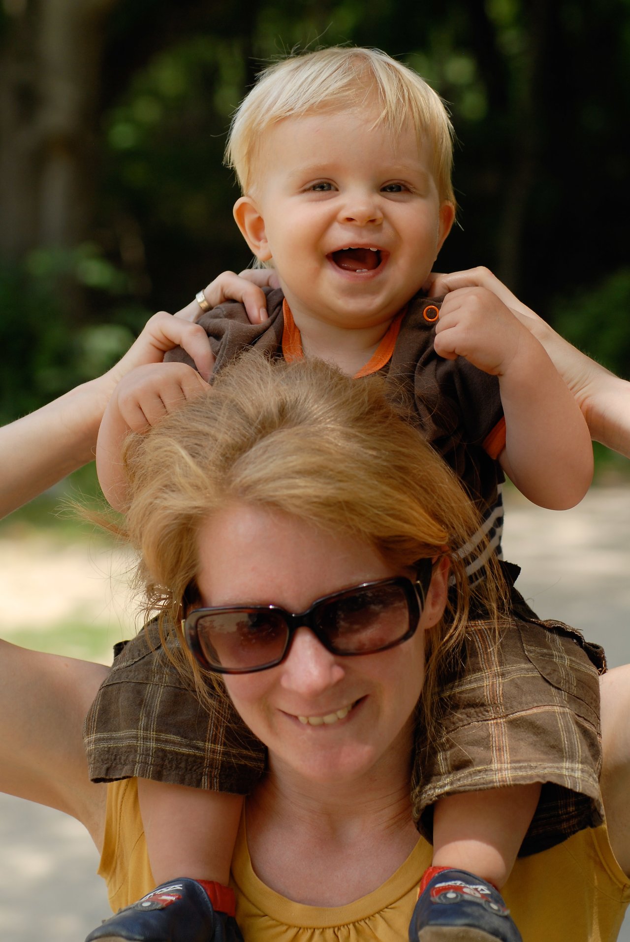 A smiling woman wearing sunglasses carries a happy baby on her shoulders while holding the baby's hands.