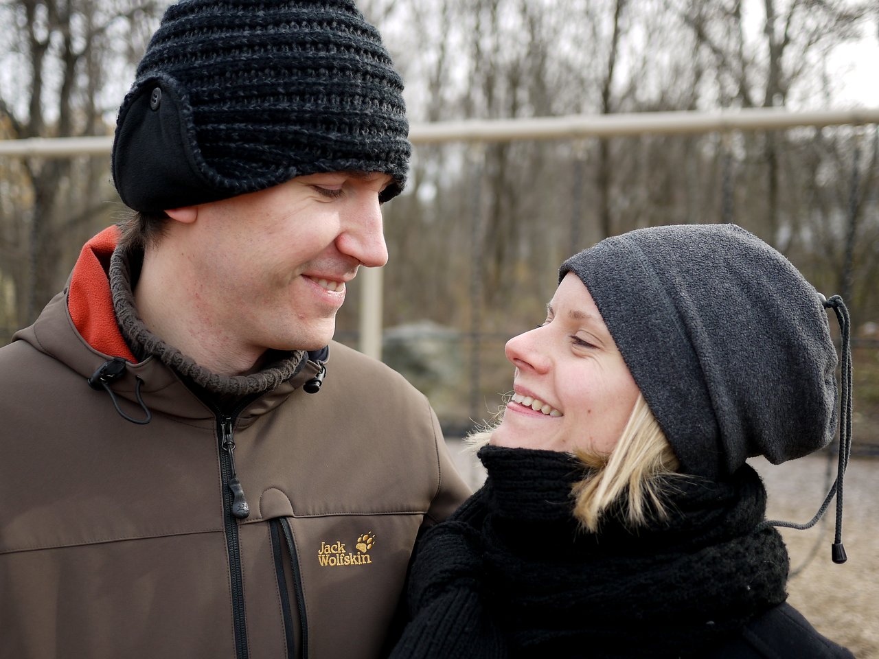 A man and woman in winter clothing smile at each other outdoors.
