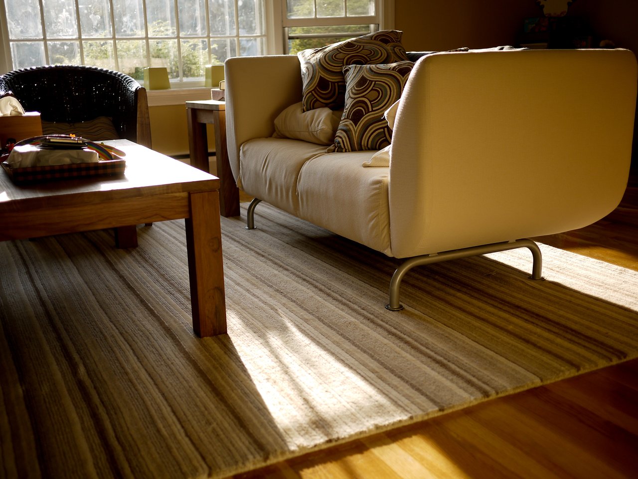 A beige sofa with patterned pillows sits on a striped rug near a wooden coffee table in a sunlit living room.