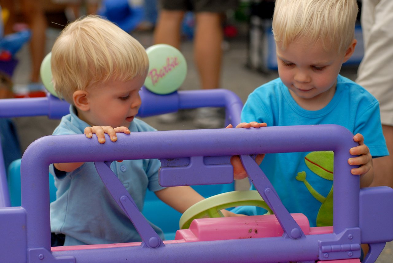 Two young children play with a pink and purple toy jeep, touching the steering wheel and dashboard.
