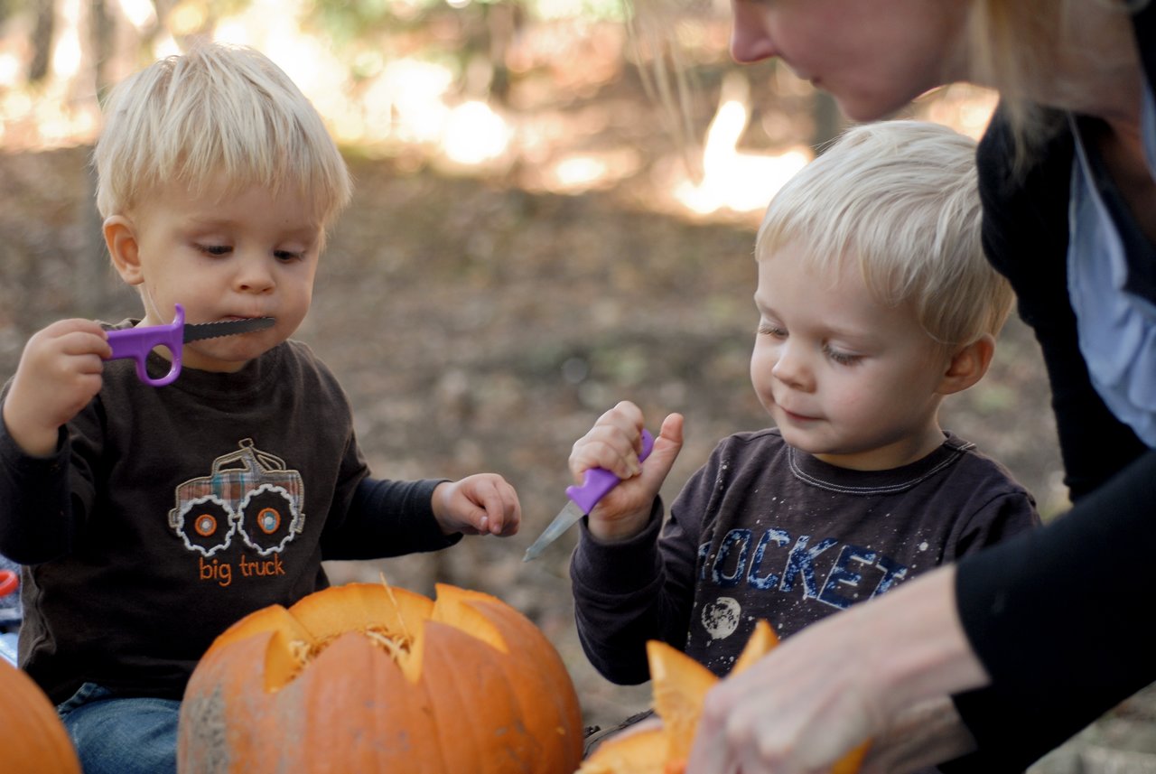 Two young children and an adult carve pumpkins outdoors, using small plastic tools to shape the designs.