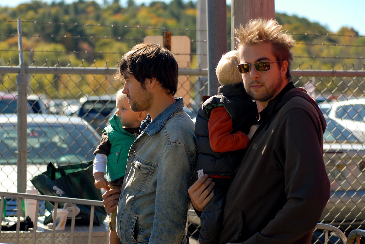 Two men stand holding young children in their arms, looking in different directions near a fenced outdoor area.