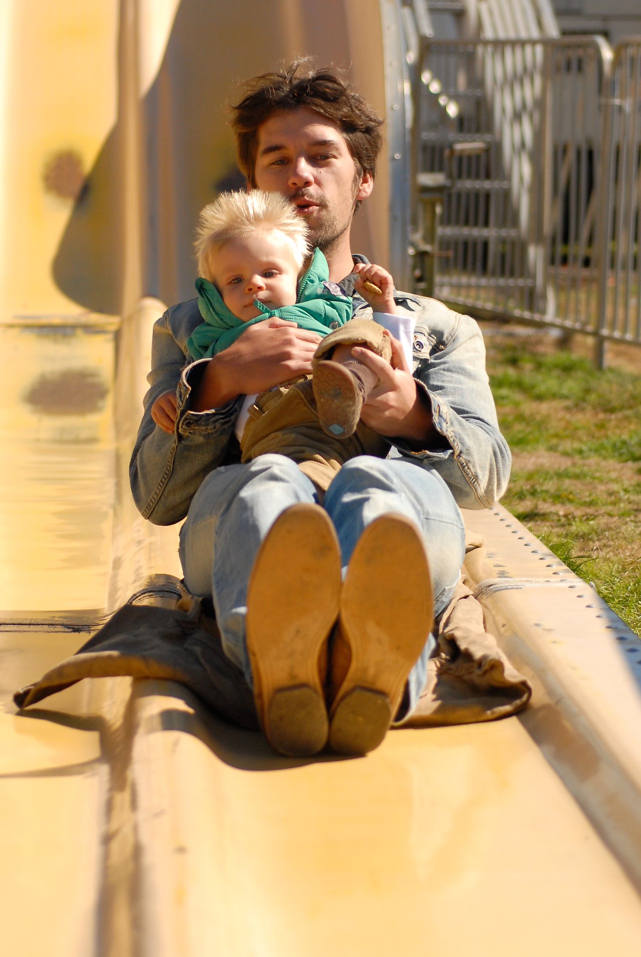 A man and a child slide down a large yellow slide together, with the child sitting on his lap.