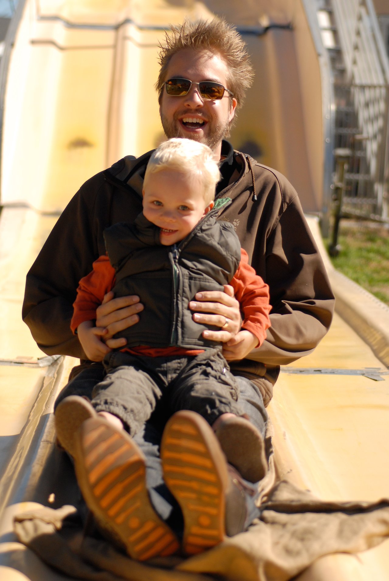 A man and a child slide down a large yellow slide together, both smiling and enjoying the ride.