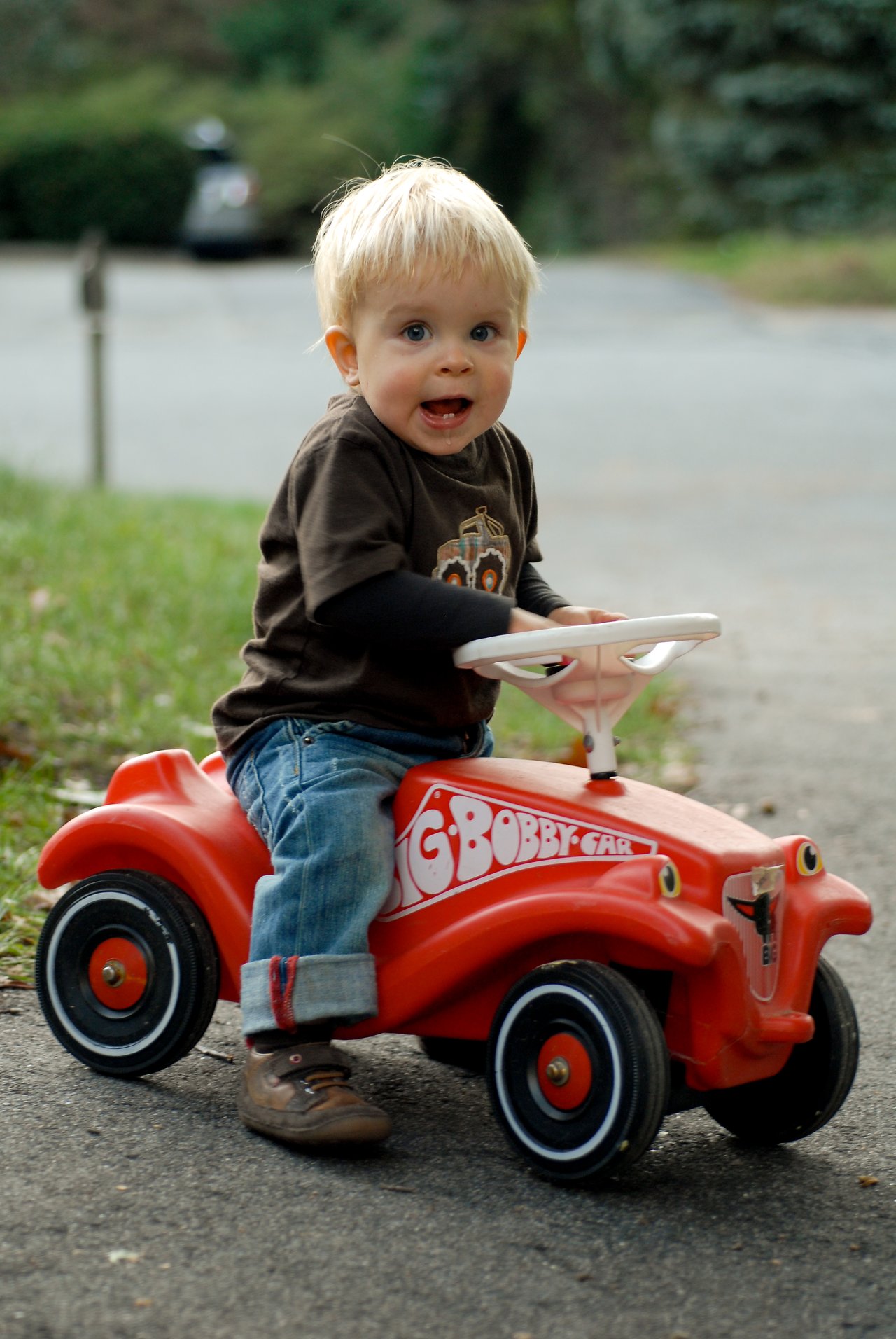 A young child sits on a red toy car, gripping the white steering wheel with an excited expression.