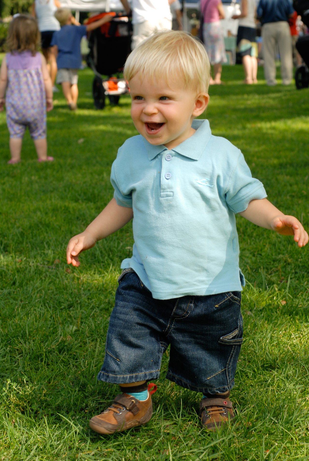 A young child with blonde hair walks on grass, smiling.