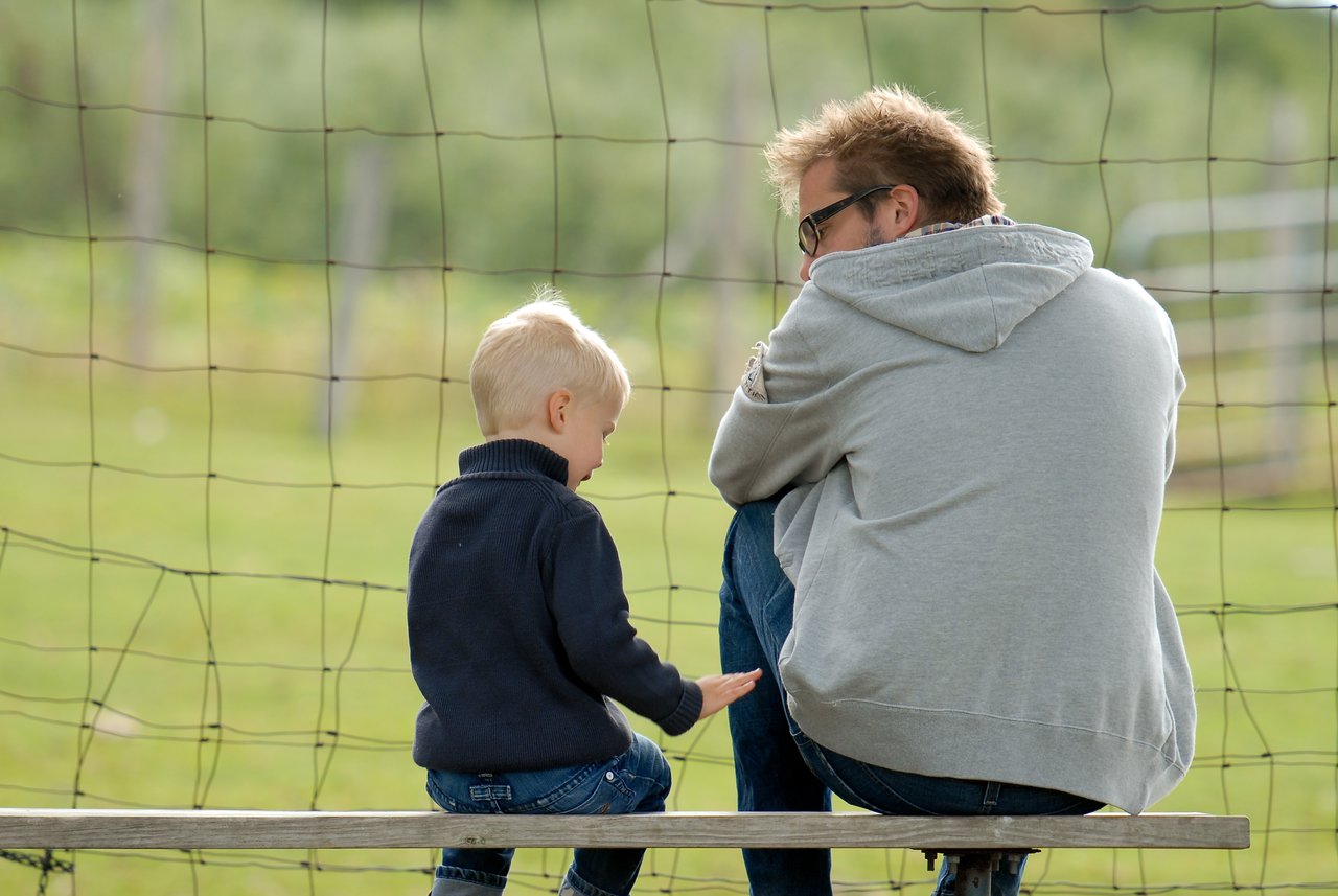 A young child and an adult sit on a bench, facing away, engaged in conversation near a netted fence.