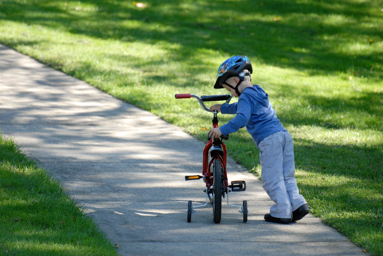 A young child wearing a helmet pushes a small red bicycle with training wheels along a paved path.