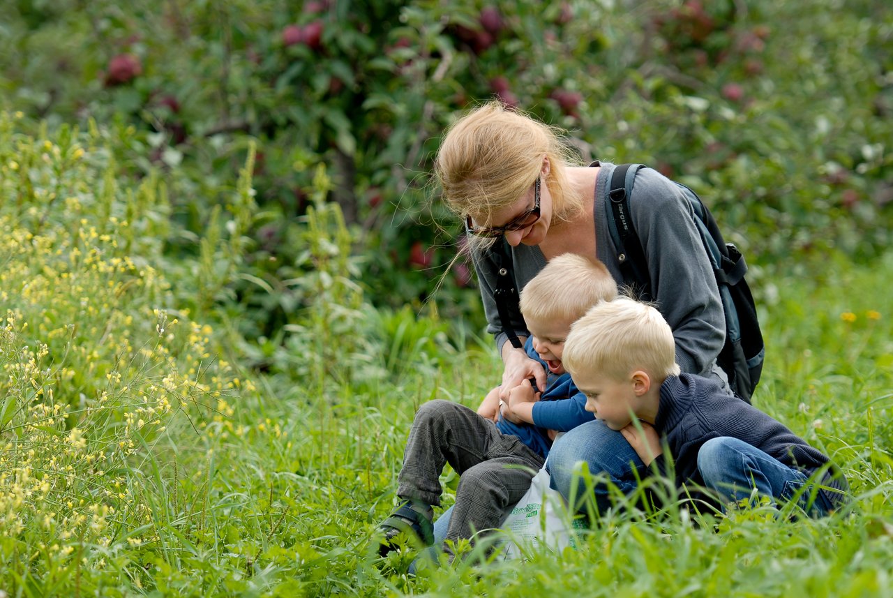 A woman crouches in the grass, playfully interacting with two young children who are laughing and smiling.