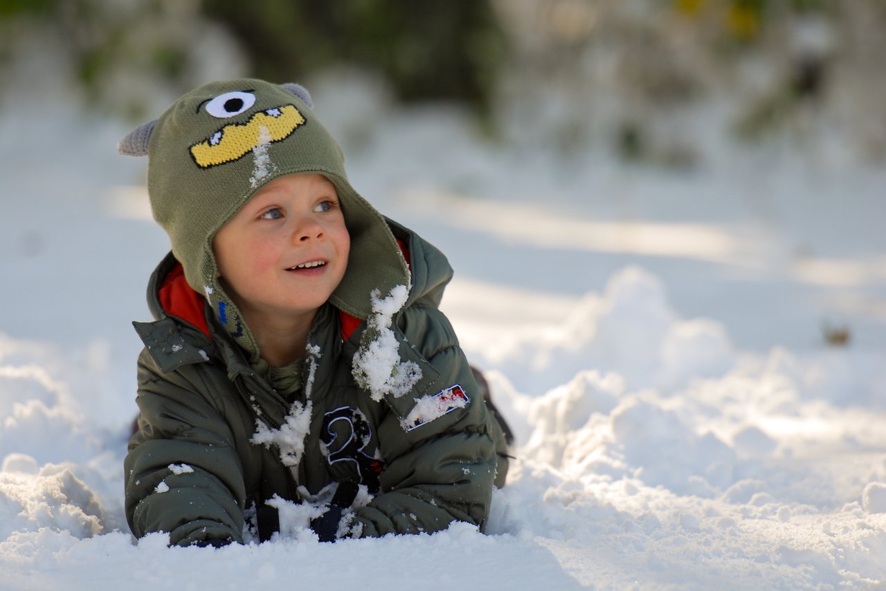 A young child in a green winter coat and monster hat lies in the snow, smiling and covered in snowflakes.