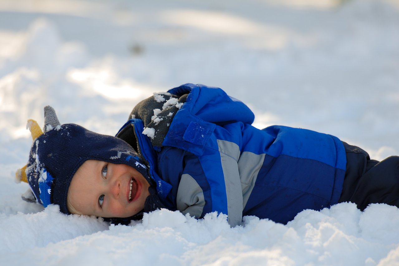 A child in a blue winter coat and hat lies in the snow, smiling and playing.