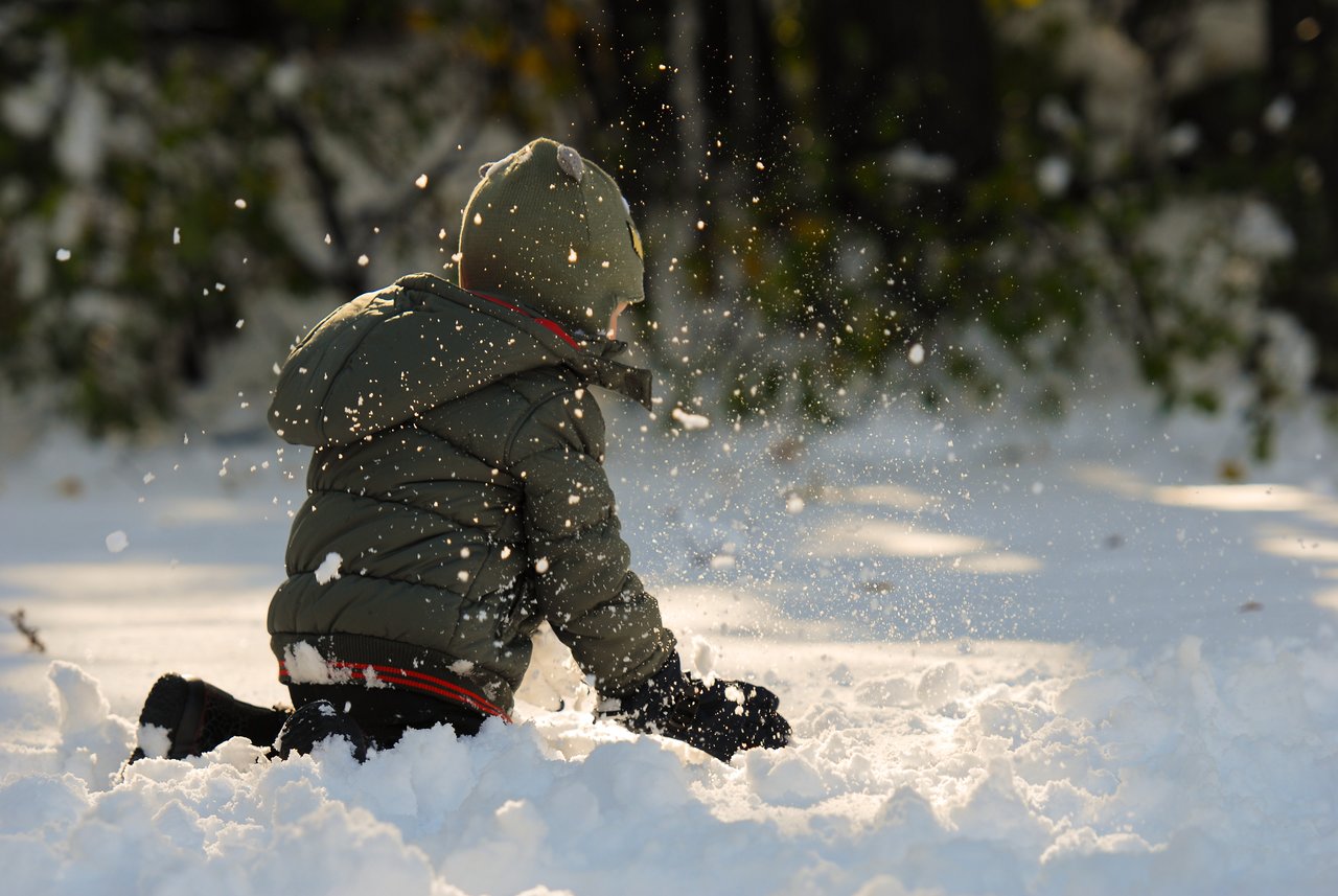 A child in a winter coat plays in the snow, tossing it into the air while kneeling.