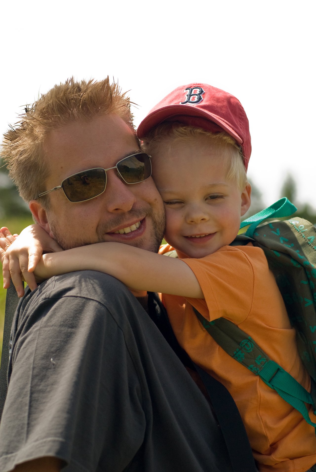 A man wearing sunglasses carries a smiling child in a red cap and backpack, who hugs him tightly.