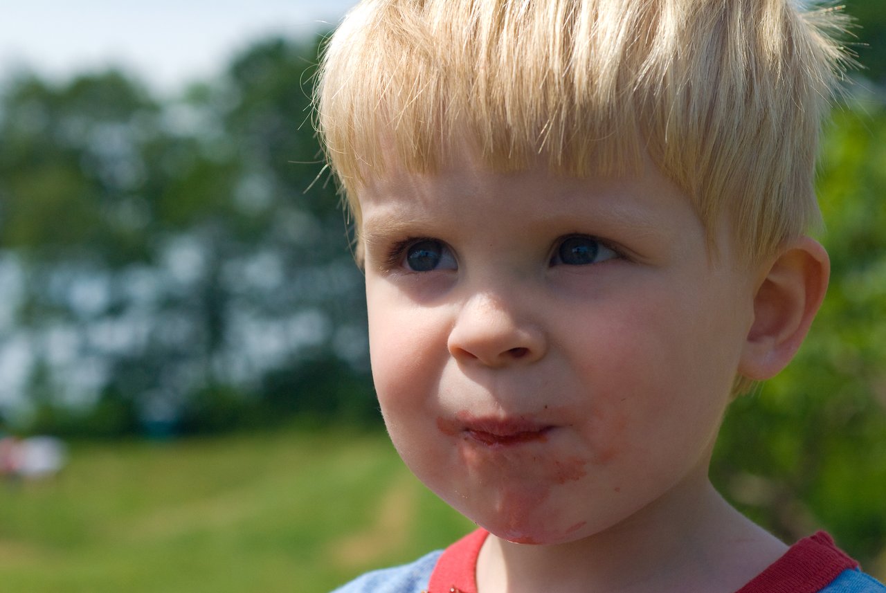 A young child with strawberry juice on their face looks off to the side while outdoors.