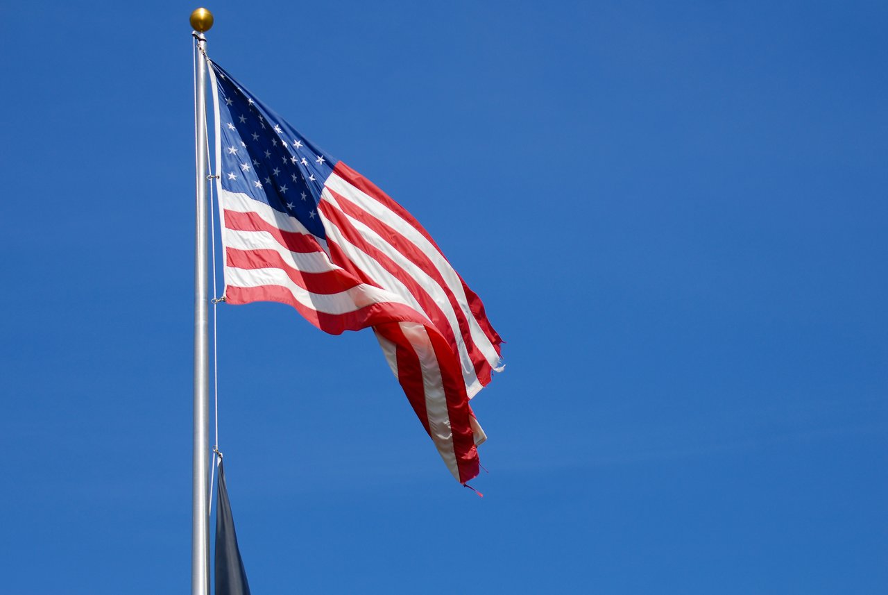 An American flag with frayed edges waves on a flagpole against a clear blue sky.