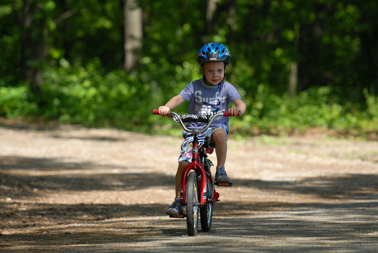 A young child wearing a helmet rides a red bicycle on a dirt path surrounded by trees.