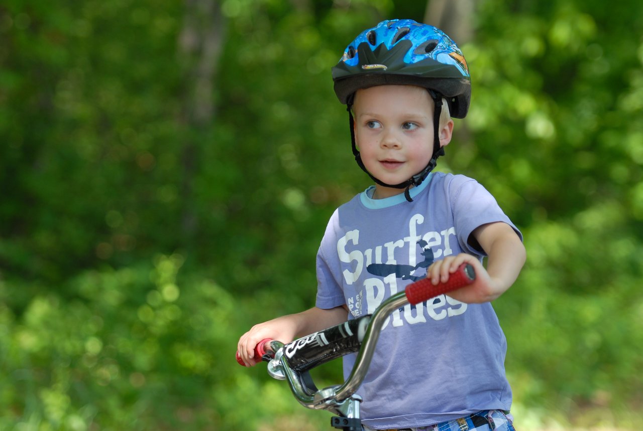 A young child wearing a helmet rides a bike on a path surrounded by green trees.