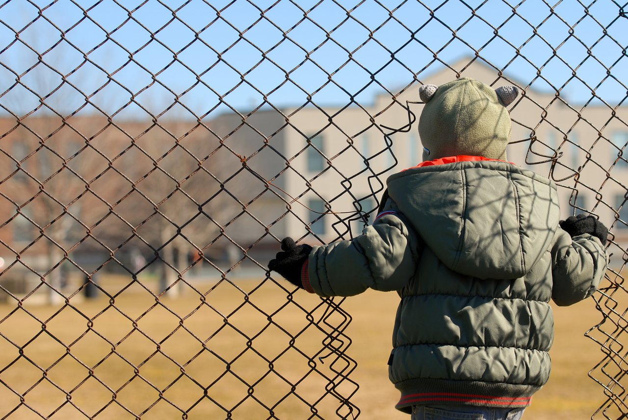 A child in a green jacket and hat pushes through a hole in a chain-link fence.