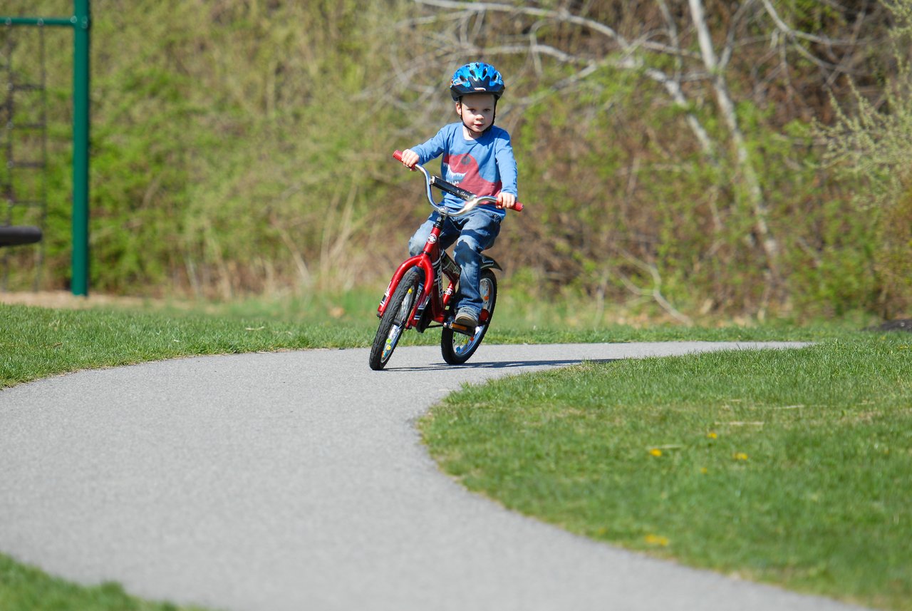 A young child rides a red bicycle without training wheels for the first time on a paved path.