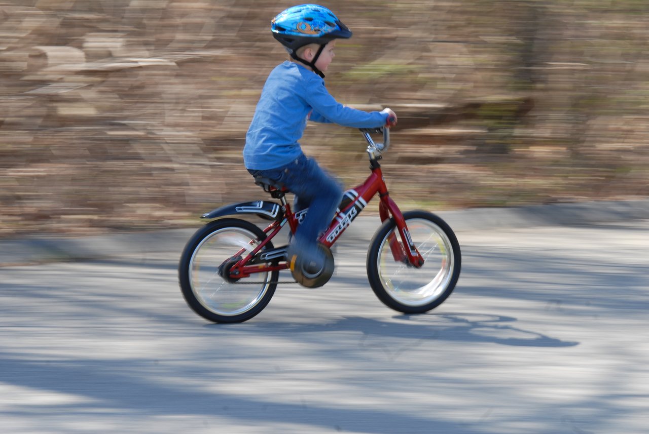 A child rides a red bicycle without training wheels for the first time, wearing a blue helmet and shirt.