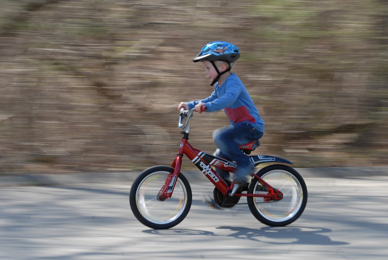 A young child wearing a helmet rides a red bicycle without training wheels for the first time.