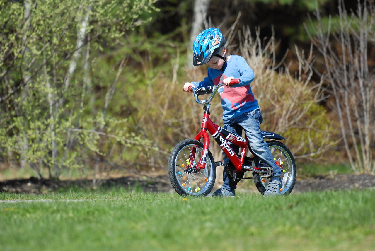 A young child wearing a helmet rides a red bicycle without training wheels for the first time.