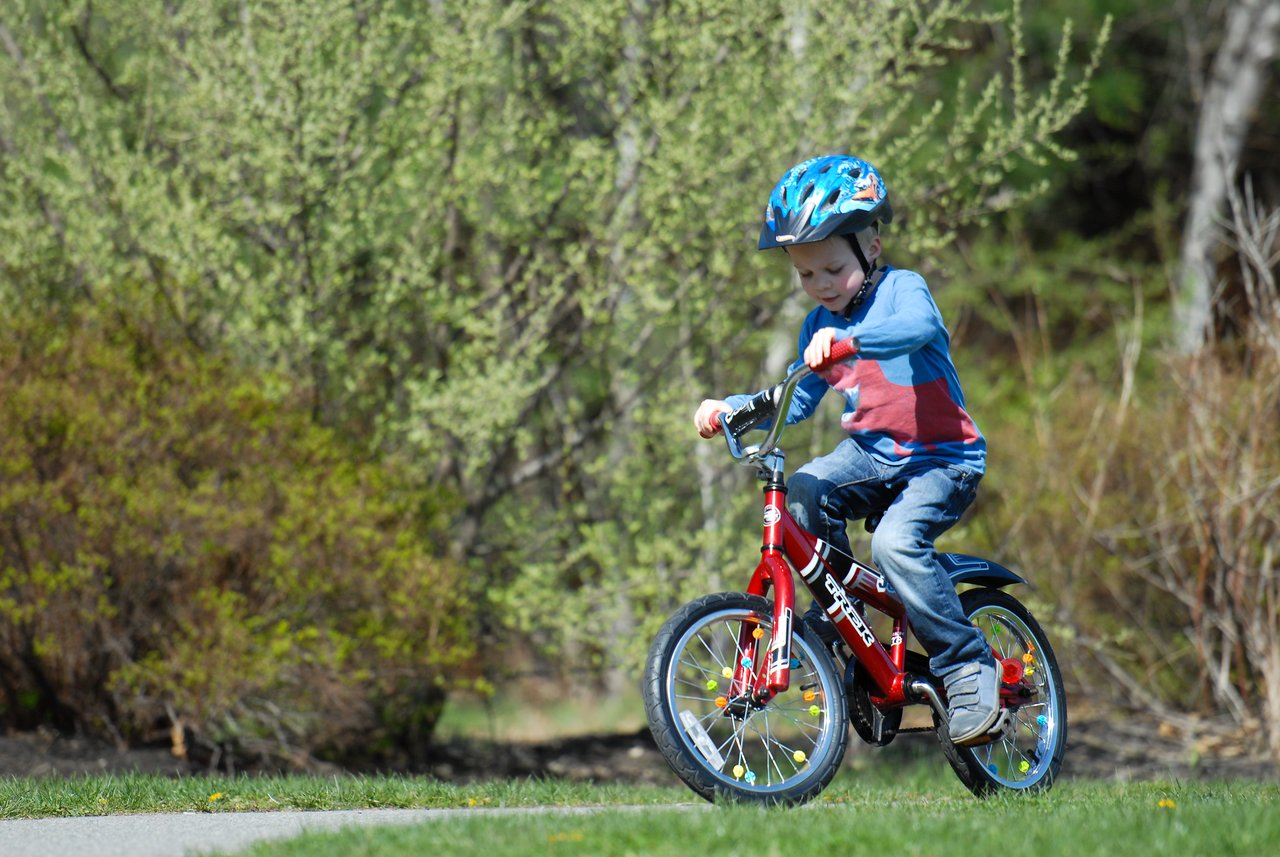 A young child rides a red bicycle without training wheels for the first time, wearing a helmet for safety.