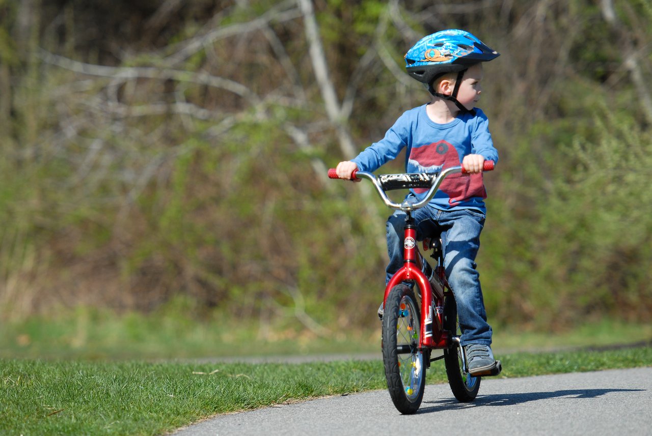 A young child wearing a helmet rides a red bicycle without training wheels for the first time.