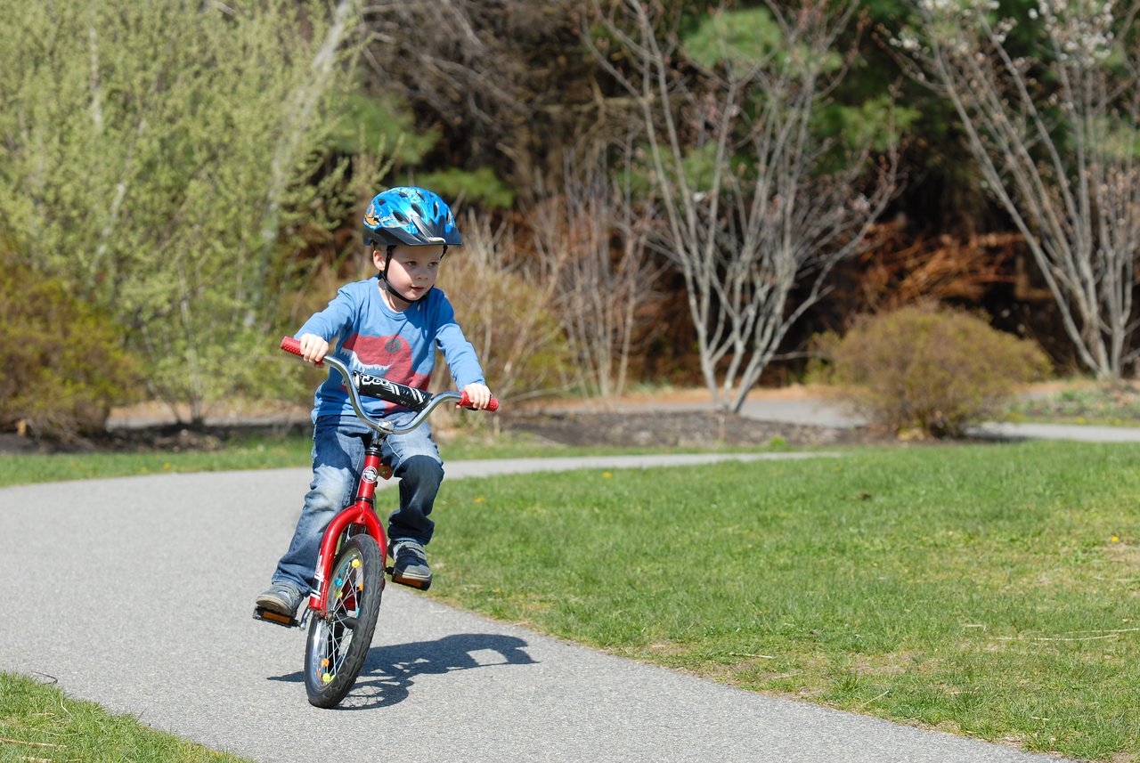 A young child rides a red bicycle without training wheels for the first time, wearing a blue helmet.