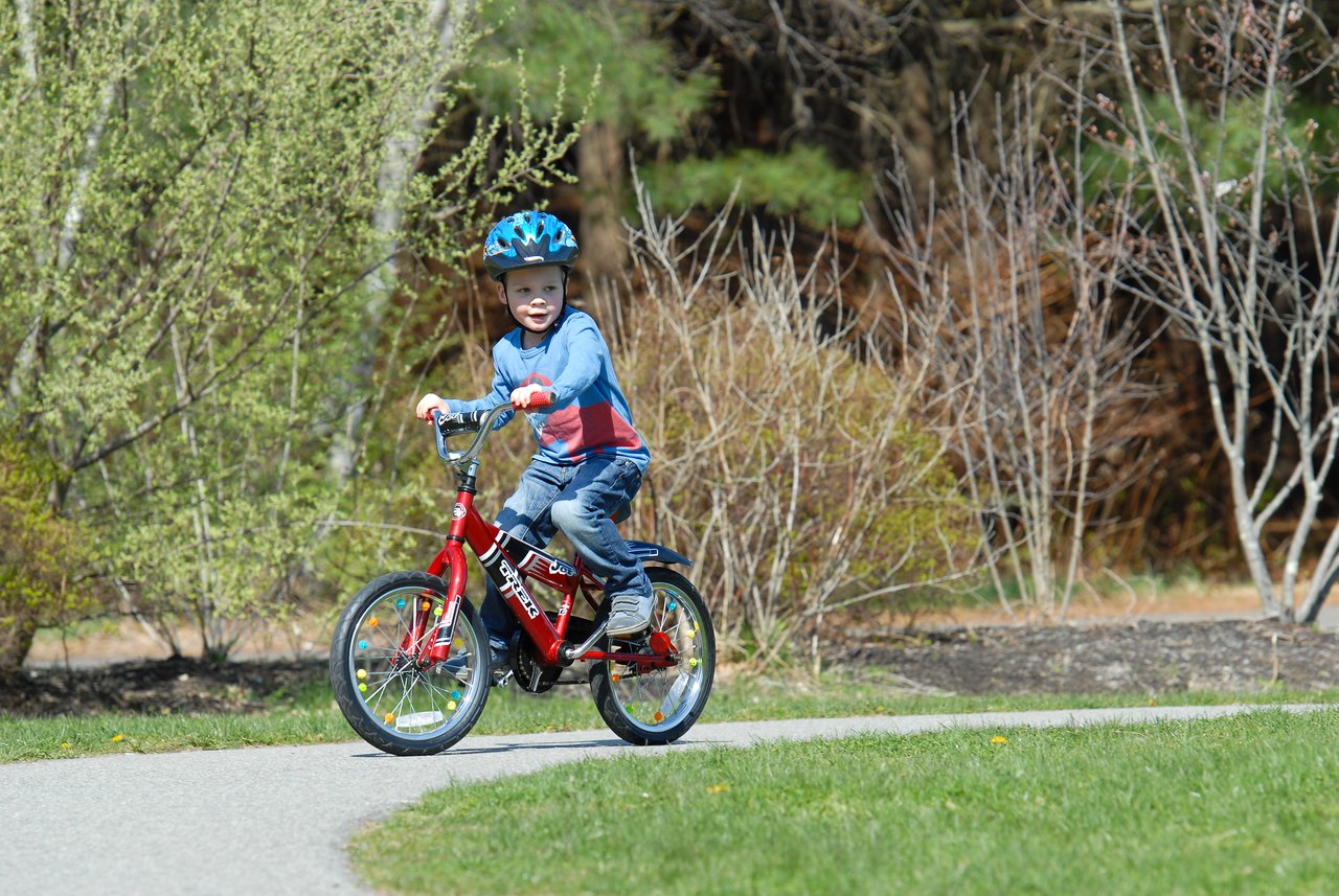 A young child rides a red bicycle without training wheels for the first time, wearing a helmet on a paved path.