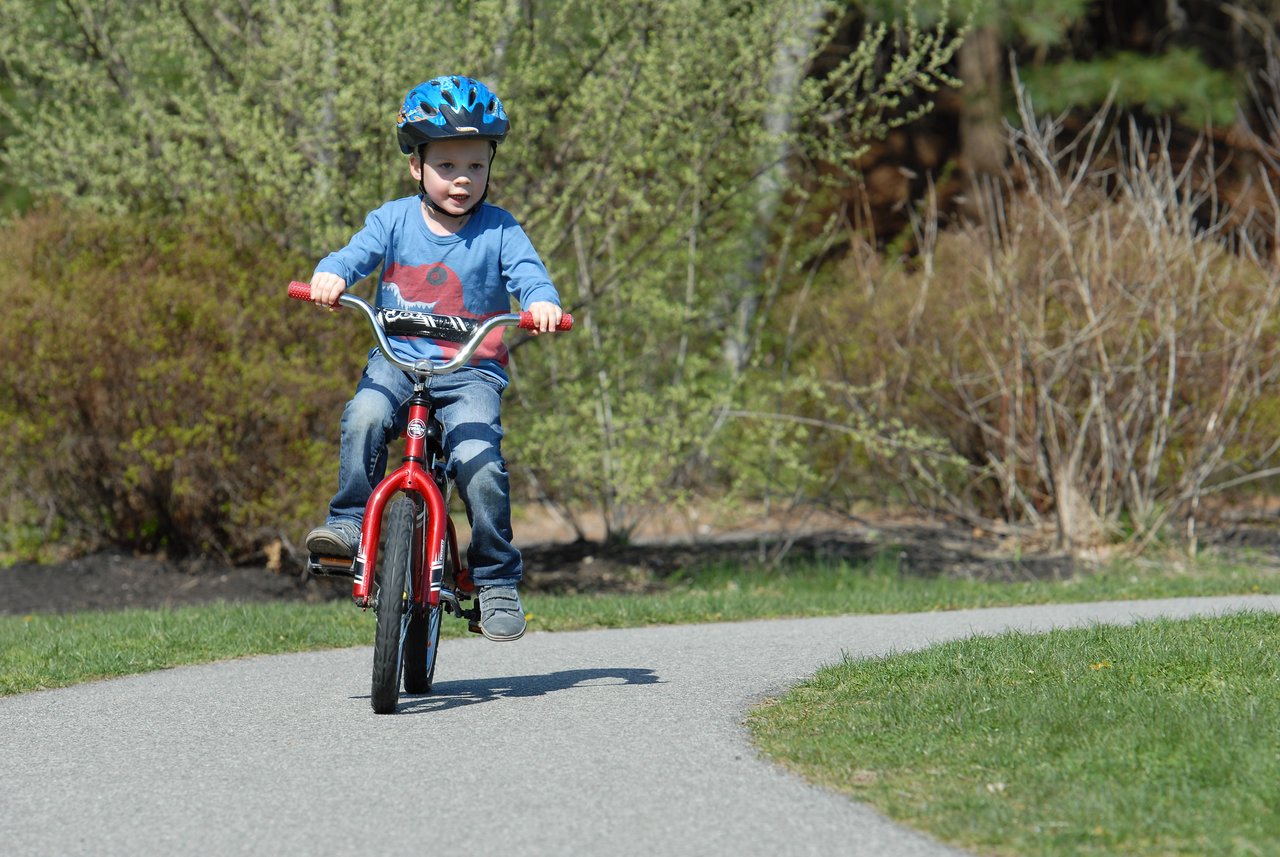 A young child rides a red bicycle without training wheels for the first time, wearing a blue helmet.