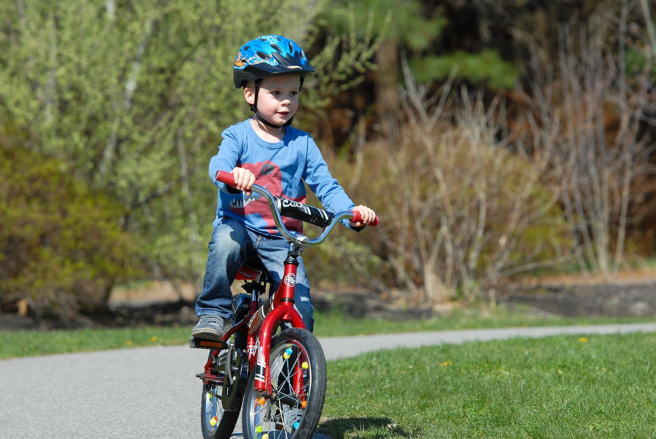 A young child rides a red bicycle without training wheels for the first time, wearing a helmet for safety.