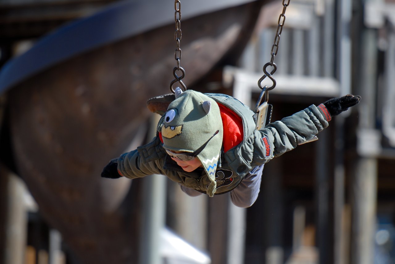 A child in a monster hat swings forward with arms outstretched, appearing to "fly" at a playground.