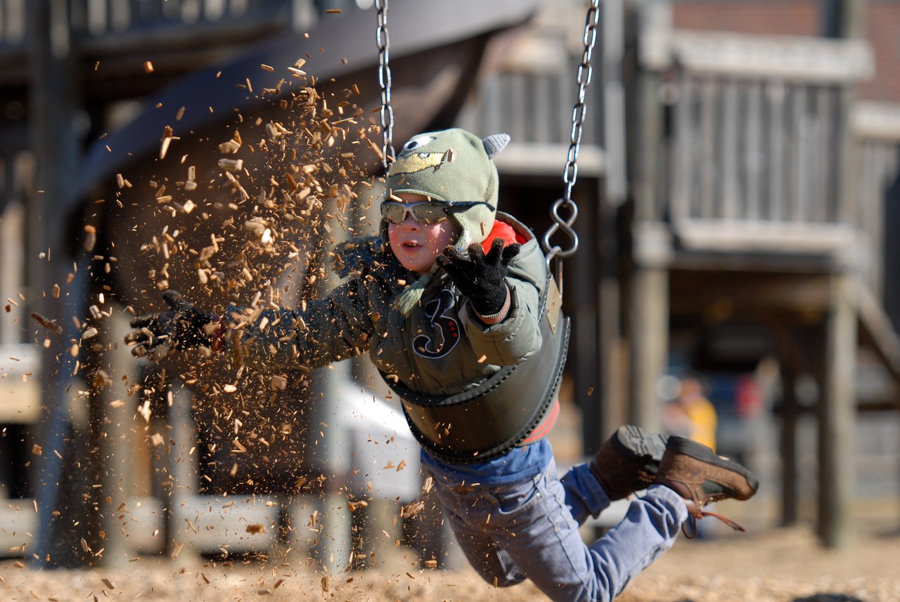 A child in a monster hat jumps off a swing, arms outstretched, kicking up wood chips in midair.