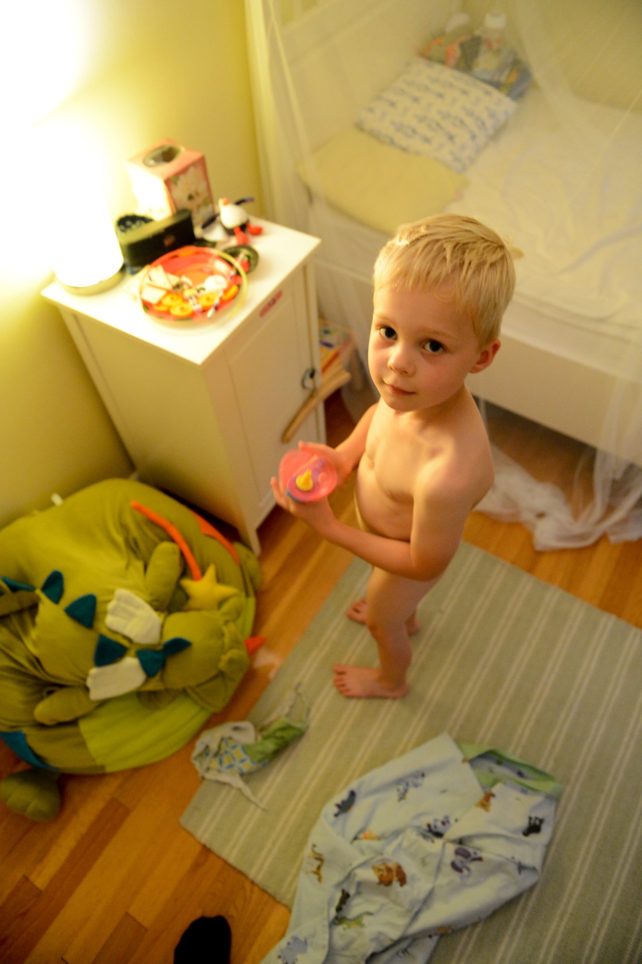 A young child stands in a bedroom holding a small cup, with pajamas on the floor, preparing for bed.