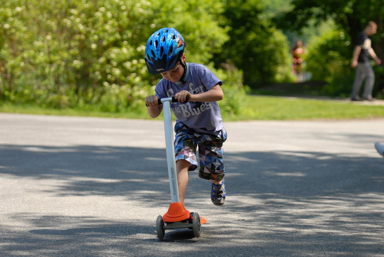 A child wearing a blue helmet rides a scooter on a paved path, pushing forward with one foot.