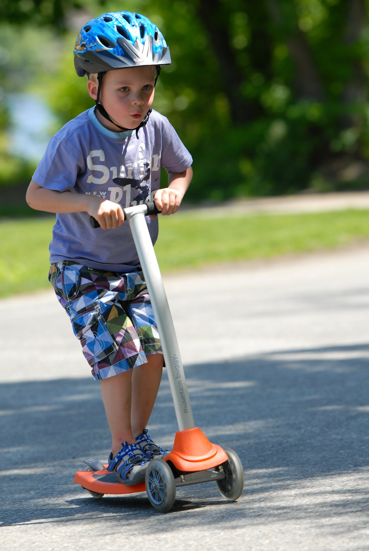A young child wearing a helmet rides a three-wheeled scooter on a paved path outdoors.
