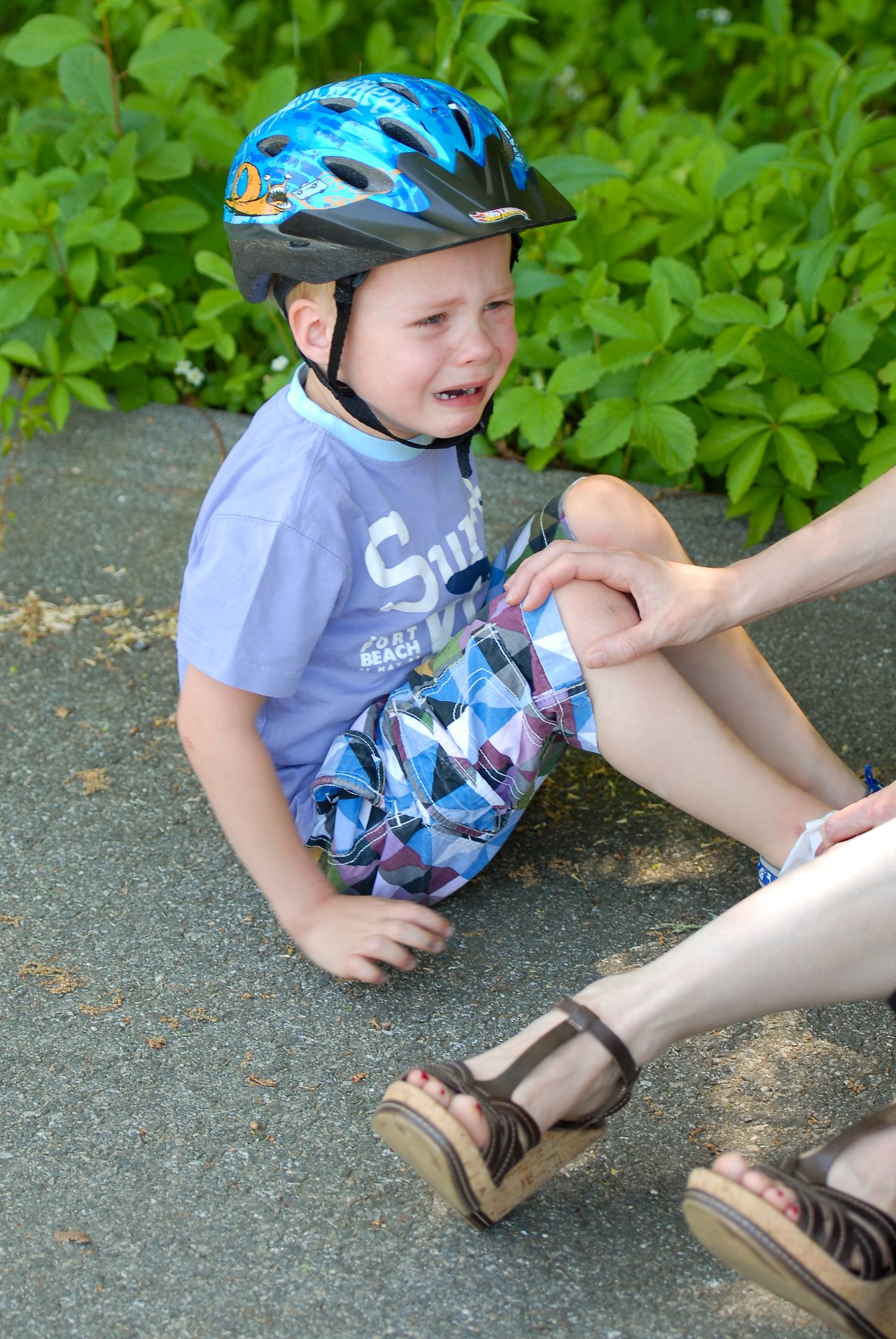 A young boy wearing a helmet sits on the ground crying while an adult comforts him.
