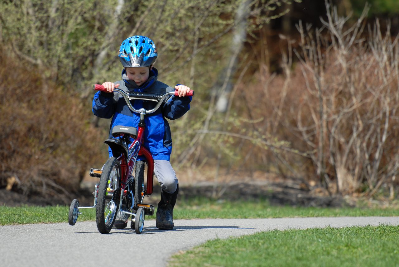 A child wearing a blue helmet and jacket walks alongside a small bike with training wheels on a paved path.