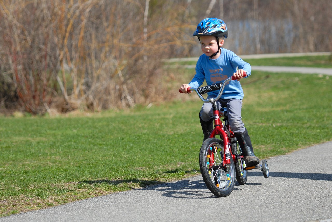 A young child wearing a helmet rides a red bicycle with training wheels on a paved path.