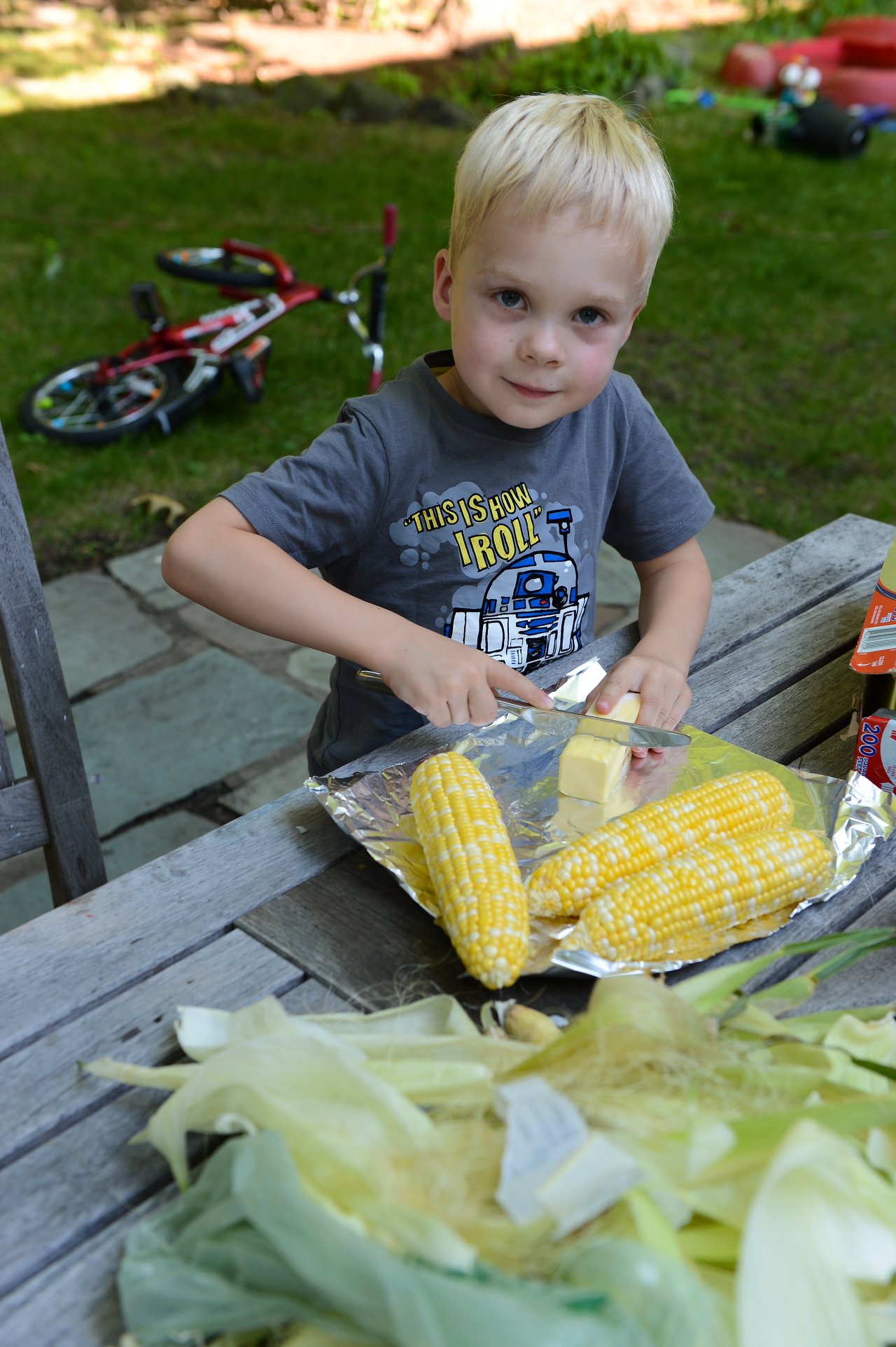 A young child spreads butter on corn while sitting at an outdoor table, preparing food in the backyard.