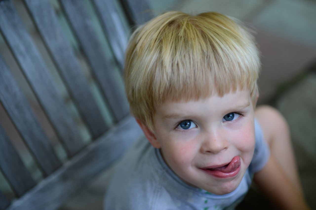 A young child with blonde hair sits outside, playfully sticking out their tongue and looking at the camera.