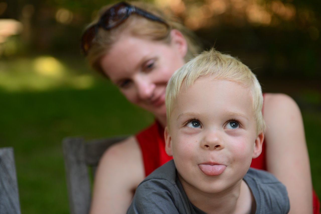 A young child sticks out their tongue playfully while an adult smiles in the background in a backyard setting.